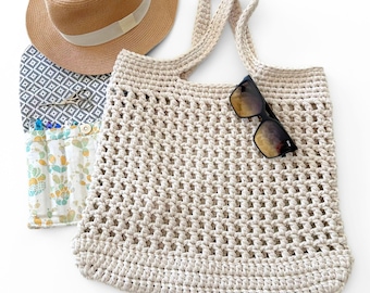 May include: A beige crocheted tote bag with an open-weave pattern and two handles, accompanied by a tan fedora hat with a white band, a pair of black sunglasses, a small pair of metal scissors, and a white pouch featuring a green cactus print.