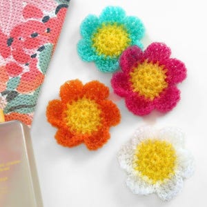 May include: Four crocheted flower scrubbers in turquoise, magenta, orange, and white, each with a yellow center. Beside them is a rectangular bottle of Native body wash with a yellow label and a floral patterned towel in shades of pink and green.