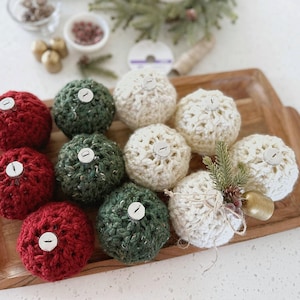 May include: A collection of round, crocheted ornaments in red, dark green, and cream colors. Each ornament features a small silver-toned cap at the top. One cream-colored ornament is decorated with a small sprig of pine and a tiny gold bell.