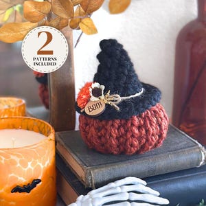 May include: A crocheted pumpkin in a dark orange yarn topped with a black crocheted witch hat. The hat is decorated with an orange flower, a small white flower, and a round wooden tag engraved with the text BOO!. A circular label reads 2 PATTERNS INCLUDED.