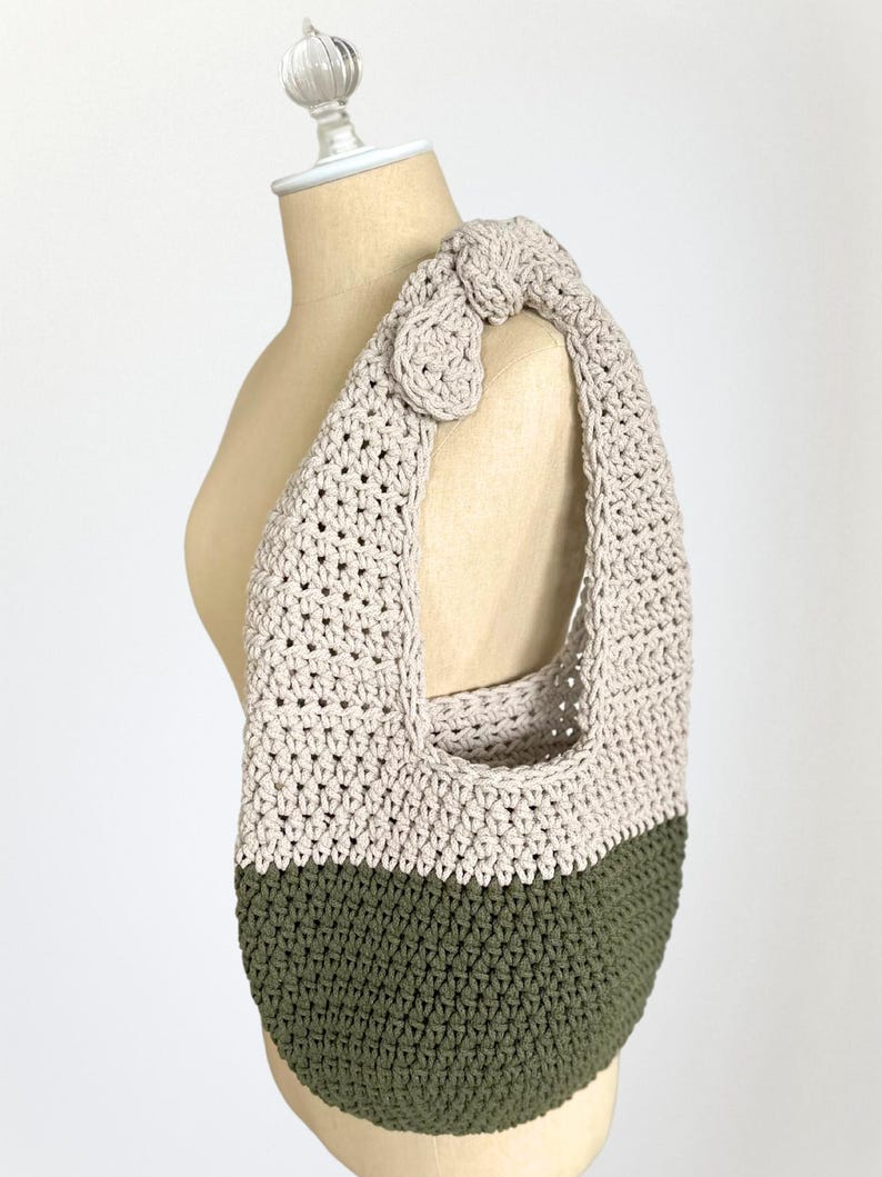 K&ouml;nnte beinhalten: Handgefertigte H&auml;keltasche mit Schulterriemen in Zweifarben-Design. Der obere Teil ist in einem neutralen Beige gehalten, w&auml;hrend der untere Teil olivgr&uuml;n ist. Der Riemen hat einen dekorativen Knoten und Knopf. Die Tasche wird auf einer B&uuml;ste pr&auml;sentiert.