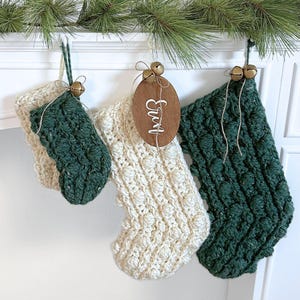 Op de afbeelding: Drie gehaakte kerstsokken met een bobbelsteekpatroon. Twee sokken zijn donker bosgroen en één is crème. Elke sok heeft een klein koperen belletje vastgebonden met touw. Een ovaal houten label met de witte inscriptie van de naam Erin is bevestigd.