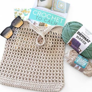 May include: A beige crochet market bag with a grid pattern, black sunglasses, a book titled Crochet to Calm, a ball of green Bernat Maker yarn, and a ball of beige I Love This Cotton yarn. The items are arranged together on a plain white surface.