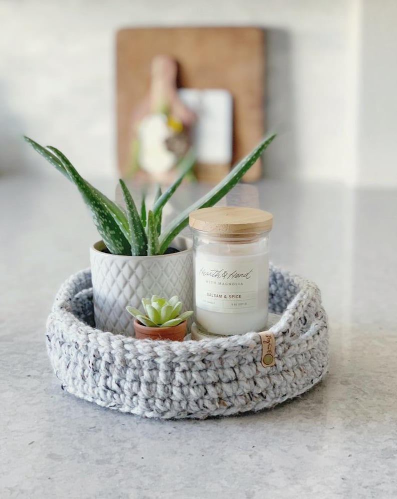 May include: A gray crocheted basket holds a small potted aloe vera plant, a succulent, and a candle in a glass jar with a wooden lid. The candle label reads "Balsam & Spice". A wooden cutting board is in the background.