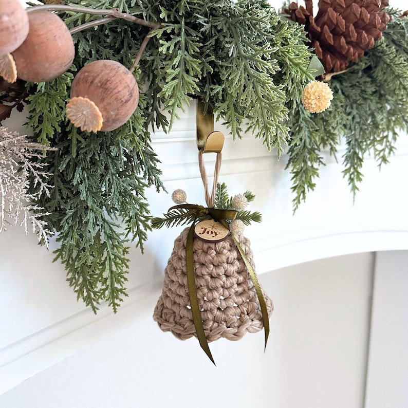 May include: A beige crocheted bell ornament hanging from a gold metal hook. The bell features a dark green ribbon tied in a bow, small white berries, and a circular wooden tag engraved with the word Joy and a small snowflake icon.