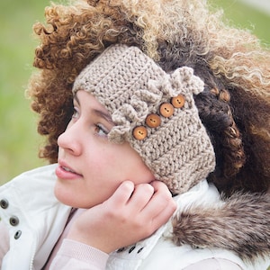 May include: A beige crochet headband featuring a textured ribbed pattern and a decorative ruffled edge on one side. The headband is accented with four small, round, light brown wooden buttons arranged in a vertical row along the side.
