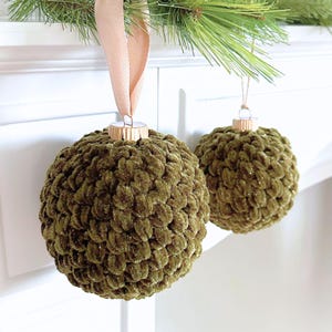 May include: Two spherical Christmas ornaments covered in olive green crocheted yarn with a subtle shimmer. Each ornament features a metallic gold cap and a soft, light pink ribbon loop for hanging, showcasing a textured, plush, and rounded design.