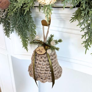 May include: A beige crocheted bell ornament featuring a dark green ribbon, a small wooden tag engraved with the word joy, and decorative sprigs of faux pine needles and white berries, suspended from a gold-colored metal hook.
