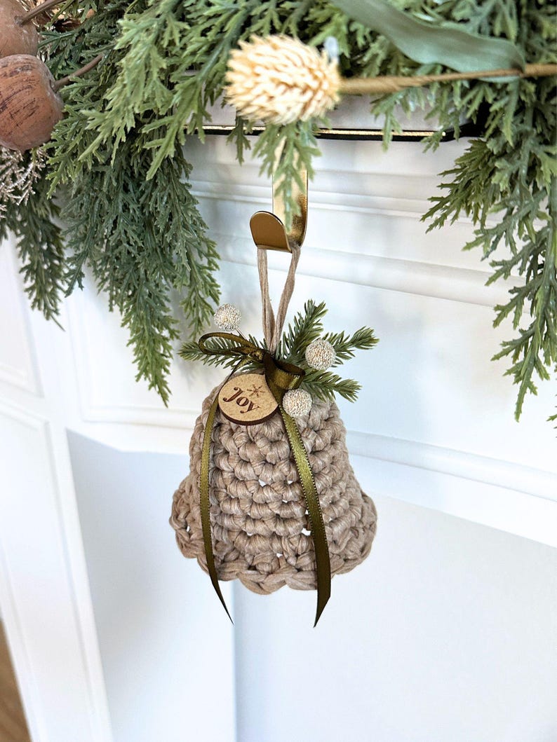 May include: A beige crocheted bell ornament featuring a dark green ribbon, a small wooden tag engraved with the word joy, and decorative sprigs of faux pine needles and white berries, suspended from a gold-colored metal hook.