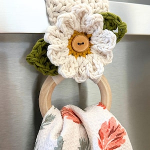 Könnte beinhalten: Ein Häkel-Handtuchhalter mit einer cremefarbenen Blume, gelber Mitte und braunem Holzknopf, flankiert von zwei grünen Häkelblättern. Ein heller Holzring hängt darunter und hält ein weißes Küchentuch mit einem floralen Druckmuster.