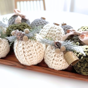 Puede incluir: Una colección de adornos redondos de hilo de ganchillo en color crema, verde oliva y gris. Cada adorno está rematado con una pequeña piña, bayas doradas brillantes y ramitas de pino escarchadas. Algunos incluyen una cinta o una pequeña campana.
