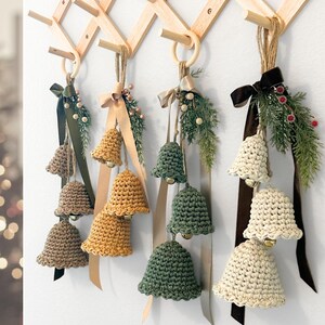 May include: Four sets of three crocheted bells in tan, mustard yellow, sage green, and cream, each hanging from a wooden ring. Each set features a satin ribbon, faux pine sprigs, and small berries, suspended from a wooden accordion wall rack.
