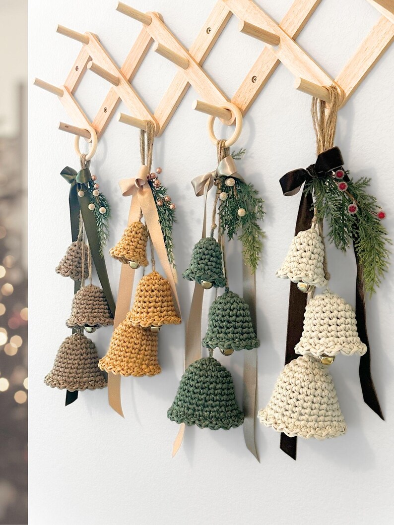 May include: Four sets of three crocheted bells in tan, mustard yellow, sage green, and cream, each hanging from a wooden ring. Each set features a satin ribbon, faux pine sprigs, and small berries, suspended from a wooden accordion wall rack.