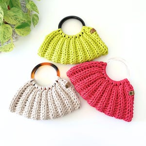 May include: Three crescent-shaped handbags made of thick, ribbed knit cord. One bag is lime green with a black circular handle, one is beige with a tortoiseshell handle, and one is hot pink with a clear circular handle. Each has a small brown tag.