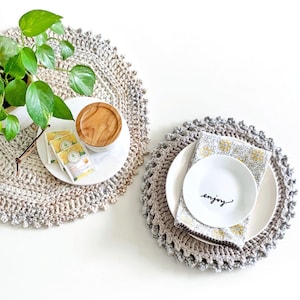 May include: Two round, textured, beige and grey crocheted placemats. One holds a white plate with a wooden-lidded jar, tea packets, and a green plant. The other holds a white plate, a floral patterned napkin, and a small white bowl with the word enjoy.