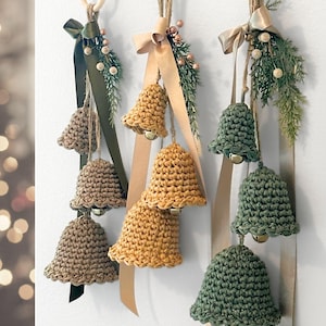 May include: Three sets of hanging crochet bells in tan, mustard yellow, and sage green. Each set features three bells of varying sizes suspended from a wooden ring by twine, accented with satin ribbons, faux pine sprigs, and small metallic berries.