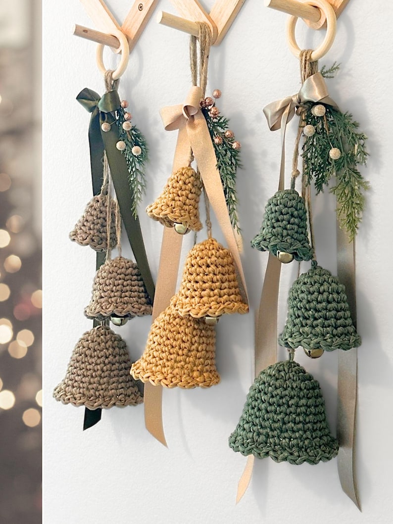 May include: Three sets of hanging crochet bells in tan, mustard yellow, and sage green. Each set features three bells of varying sizes suspended from a wooden ring by twine, accented with satin ribbons, faux pine sprigs, and small metallic berries.