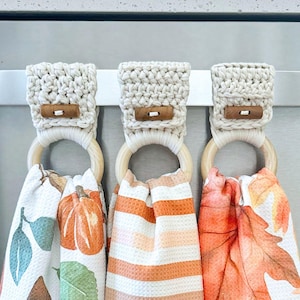 May include: Three hanging kitchen towels with crochet tops in cream yarn, each secured with a wooden toggle button and a light-colored wooden ring. The towels feature patterns of orange pumpkins, horizontal stripes, and autumn leaves in warm tones.