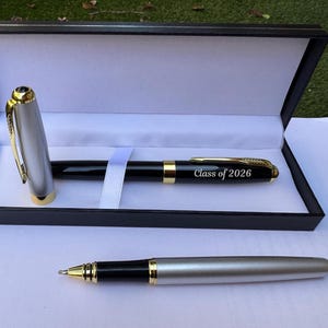Pode incluir: Uma caneta preta e prateada com detalhes dourados, apresentada numa caixa preta com interior branco. A caneta está gravada com "Class of 2026". Uma segunda caneta é visível abaixo. Um ótimo presente de formatura.