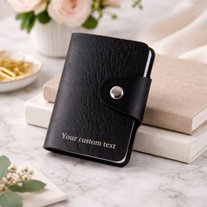 Puede incluir: Tarjetero de cuero negro con cierre de botón a presión plateado. El texto "Your custom text" está impreso en la parte delantera. El tarjetero está sobre un libro y una superficie blanca.