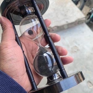 May include: Black hourglass with clear glass bulbs and black sand. The hourglass has a dark wooden frame and base. The hourglass is held in a hand, with the sand flowing from the top bulb to the bottom bulb.