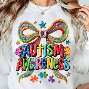 May include: White sweatshirt featuring a colorful design promoting autism awareness. The design includes a rainbow-colored bow, puzzle pieces, stars, and the words "AUTISM AWARENESS" in vibrant, multi-colored letters.