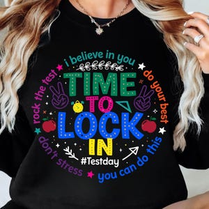 Könnte beinhalten: Schwarzes Sweatshirt mit einem farbenfrohen Aufdruck, der "Time to Lock In" zeigt, zusammen mit den Worten "I believe in you", "Rock the test", "Do your best", "Don't stress" und "You can do this". Das Design beinhaltet ein Peace-Zeichen, einen Apfel und eine Uhr.