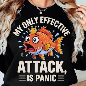 May include: Black sweatshirt with the text "MY ONLY EFFECTIVE ATTACK, IS PANIC" and a cartoon goldfish wearing a crown. The goldfish is orange with yellow fins and a surprised expression. Water droplets surround the fish.