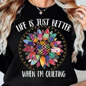 May include: Black sweatshirt with the text "LIFE IS JUST BETTER WHEN I'M QUILTING." The design features a colourful sunflower made of patchwork fabric. The sunflower is surrounded by a gold circle with star accents.