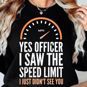 Op de afbeelding: Zwart sweatshirt met de tekst "YES OFFICER I SAW THE SPEED LIMIT I JUST DIDN'T SEE YOU". Boven de tekst staat een oranje snelheidsmeter met de letters "MPH".