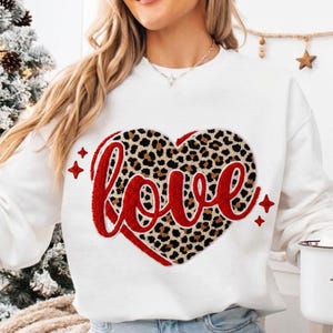 Puede incluir: Sudadera blanca con un diseño de corazón con estampado de leopardo y la palabra "love" en letras rojas. Acentos de estrellas rojas rodean el corazón. Una prenda casual y cómoda.