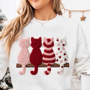 May include: White sweatshirt featuring four embroidered cats in pink, red, and white, perched on a brown branch. The cats have detailed textures, with one cat adorned with heart-shaped accents. A cozy and charming design.