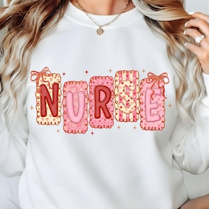 Puede incluir: Sudadera blanca con la palabra "NURSE" en un diseño de San Valentín. Las letras son rosas y rojas, decoradas con corazones y espirales. Cada letra está enmarcada con un borde festoneado y un lazo.