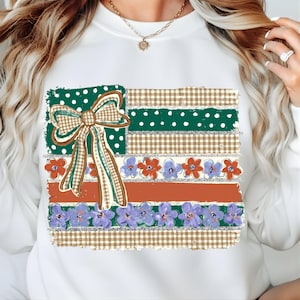 Può includere: Felpa bianca con un design grafico colorato. Il design include una sezione a pois verdi, un fiocco a quadri marrone e verde e strisce di vichy marrone, arancione e una fila di fiori viola e rossi. La felpa ha uno scollo tondo.