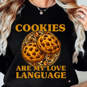 May include: Black sweatshirt with the text "COOKIES ARE MY LOVE LANGUAGE" in gold lettering. The design features two chocolate chip cookies in a decorative gold circle.