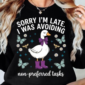 Puede incluir: Sudadera negra con el texto "SORRY I'M LATE. I WAS AVOIDING non-preferred tasks." Un ganso blanco con un lazo y botas moradas está rodeado de mariposas, abejas y flores.