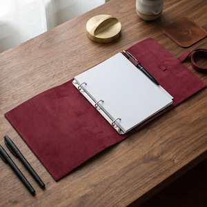Peut inclure: Un carnet en cuir bordeaux avec un classeur à trois anneaux, ouvert sur un bureau en bois. Un stylo noir repose sur les pages ouvertes. Deux stylos noirs supplémentaires sont à gauche. Un accessoire de bureau doré et une petite plante sont également sur le bureau.