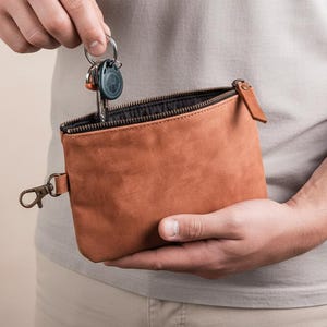 May include: A brown leather pouch with a zipper closure and a small loop on the side. The pouch is held open, revealing keys inside. A metal clasp is attached to the side. The pouch is approximately 6 inches wide and 4 inches tall.
