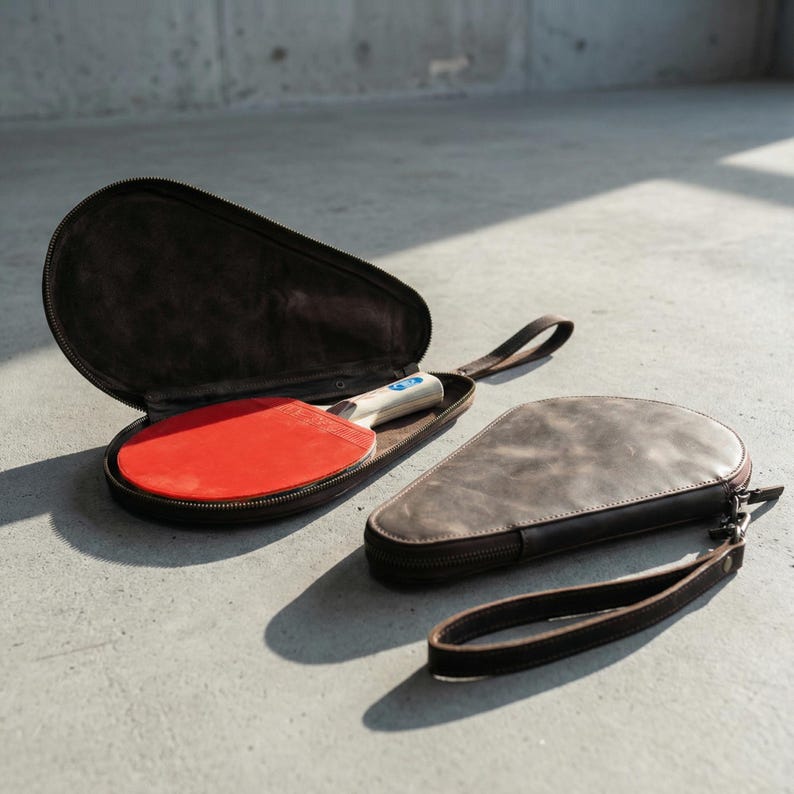 May include: Two brown leather ping pong paddle cases, one open with a red paddle inside. The other case is closed with a zipper and a wrist strap. The cases are on a concrete surface.