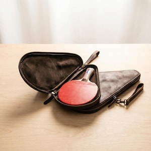 May include: A brown leather ping pong paddle case with a matching paddle inside. The paddle has a red rubber surface and a light-colored wooden handle. The case is open, revealing the paddle, and has a zipper closure and a wrist strap.