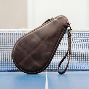 May include: A brown leather table tennis paddle case with a zipper closure and a wrist strap. The case is shaped to fit a paddle and is resting on a blue table with a net.