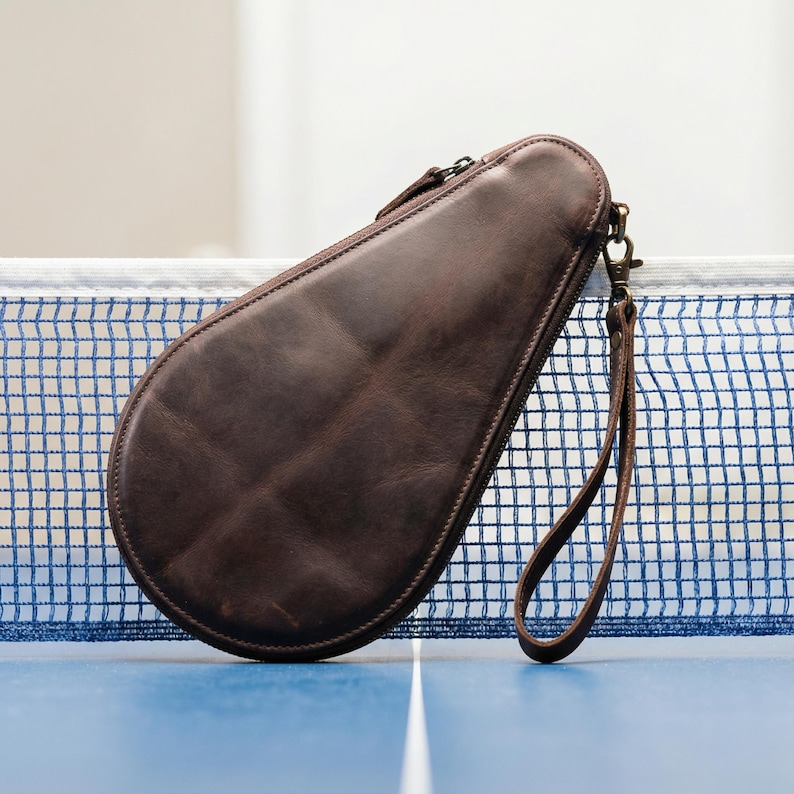 May include: A brown leather table tennis paddle case with a zipper closure and a wrist strap. The case is shaped to fit a paddle and is resting on a blue table with a net.