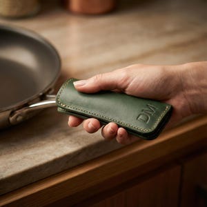 Può includere: Una protezione per il manico della padella in pelle verde scuro con le iniziali "DM" in rilievo sul davanti. La copertura è cucita con filo di colore chiaro e viene tenuta vicino a una padella in acciaio inossidabile.