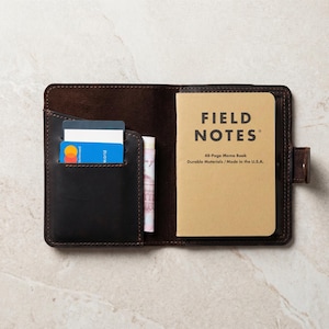 May include: Dark brown leather wallet, open to reveal a pocket with credit cards and a cash slot. A Field Notes memo book is on the right. The book is tan with black text that reads "FIELD NOTES".