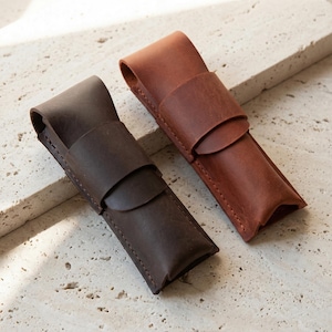 Puede incluir: Dos estuches de cuero para bolígrafos en tonos marrones. Uno es de color chocolate oscuro y el otro es de un marrón más claro, rojizo. Cada estuche tiene un cierre de solapa y un diseño cosido, lo que sugiere artesanía hecha a mano. Los estuches se muestran sobre una superficie texturizada.