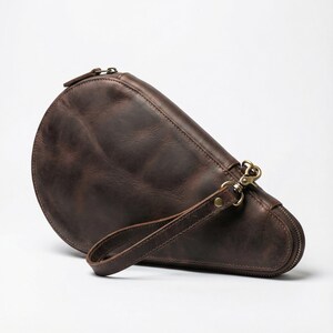 May include: A brown leather clutch with a semi-circular design. The clutch features a zipper closure, a wrist strap, and brass-colored hardware. The leather has a distressed appearance.