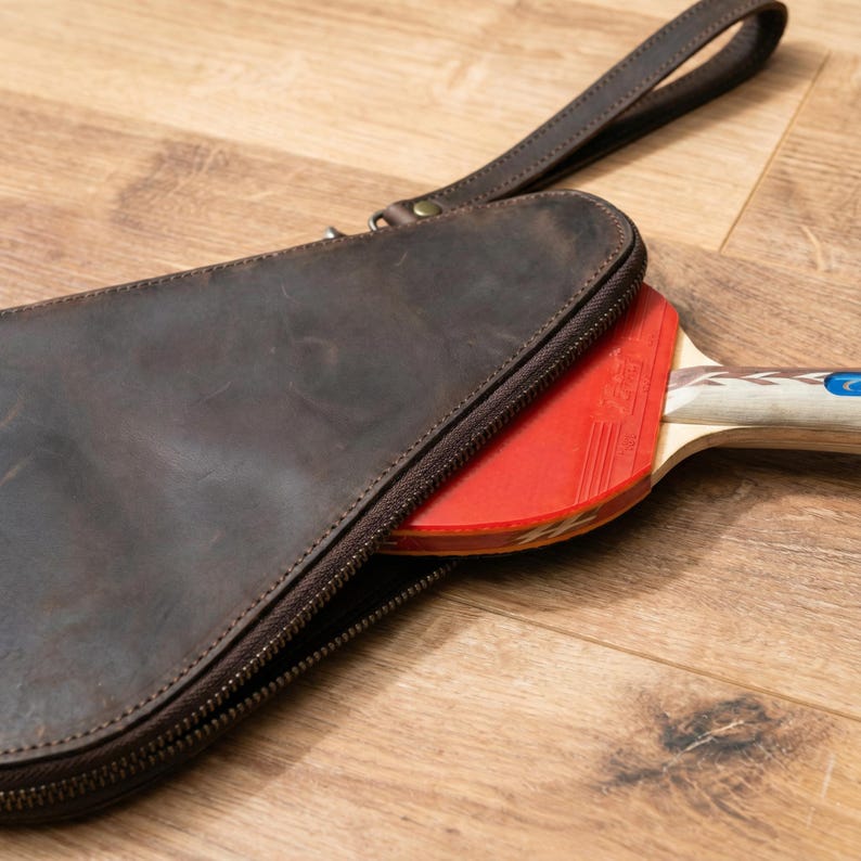 May include: A brown leather ping pong paddle case with a zipper and handle. A red and black ping pong paddle is partially visible inside the case. The case is on a wooden surface.