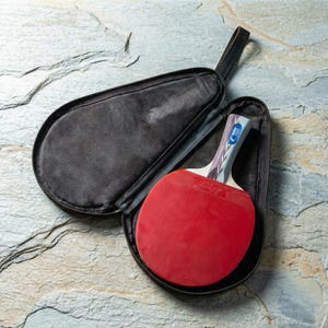 May include: A red ping pong paddle with a patterned handle rests inside an open black zippered case. The paddle has a red rubber surface and a blue and white design on the handle. The case has a black handle.