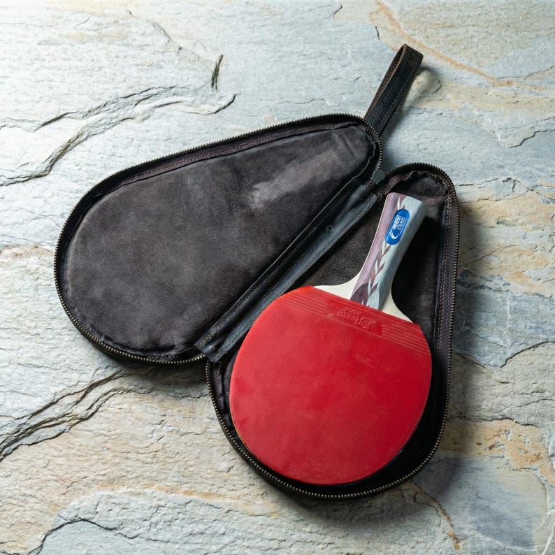 May include: A red ping pong paddle with a patterned handle rests inside an open black zippered case. The paddle has a red rubber surface and a blue and white design on the handle. The case has a black handle.