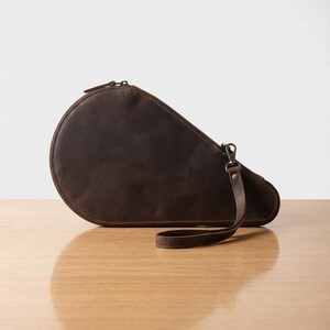 May include: A brown leather clutch with a rounded top and a zipper closure. The clutch has a wrist strap attached with a metal clasp. The bag is sitting on a wooden surface.
